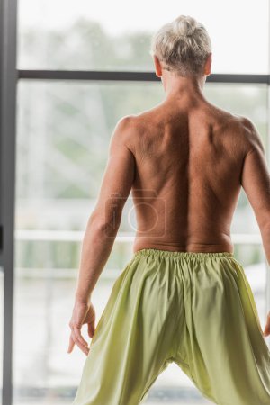back view of shirtless man in green pants standing in yoga studio 