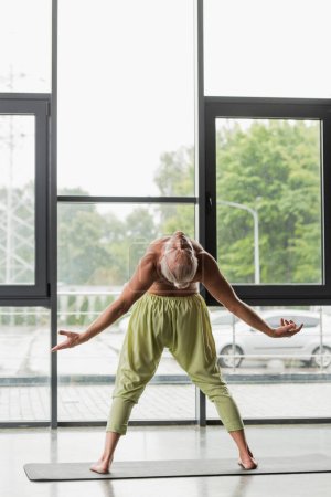 Téléchargez les photos : Pleine longueur de l'homme torse nu en pantalon vert faisant plier le dos dans le studio de yoga - en image libre de droit