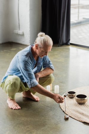 Photo for Grey haired man burning incense stick near Tibetan singing bowls and candles in yoga studio - Royalty Free Image