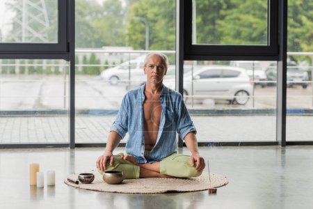 Grauhaariger Mann meditiert in der Nähe tibetischer Klangschalen und Räucherstäbchen im Yoga-Studio 