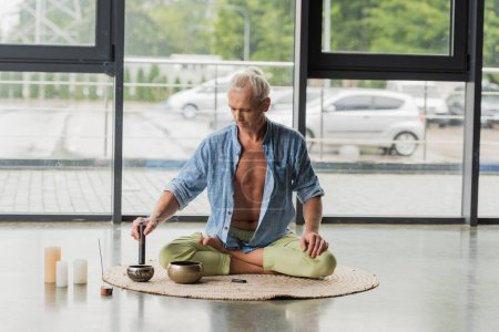 hombre de pelo gris sentado en pose de loto mientras juega en el cuenco tibetano en el estudio de yoga 