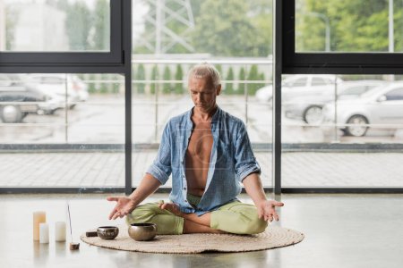 siwe włosy mężczyzna siedzi w pozycji lotosu podczas medytacji w pobliżu tybetańskiego śpiewu miski w studio jogi 
