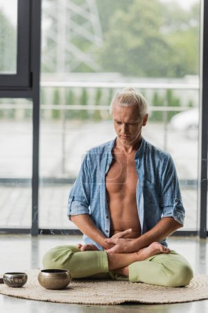 hombre sentado en pose de loto mientras medita cerca de cuencos de canto tibetano en estudio de yoga