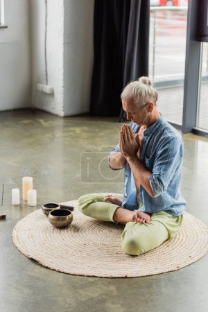 grey haired man meditating with praying hands near Tibetan singing bowls and incense stick in yoga studio 