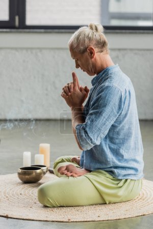 widok z boku siwy mężczyzna medytujący w pobliżu tybetańskich miski śpiewu i świece w studio jogi 
