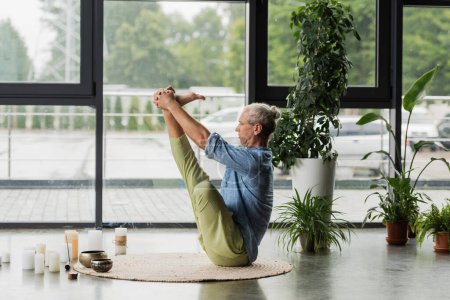 Photo for Grey haired man doing boat yoga pose near Tibetan singing bowls and incense stick in yoga studio - Royalty Free Image