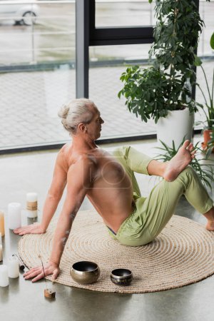 side view of grey haired man stretching back in yoga pose near candles and Tibetan singing bowls in studio 