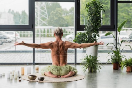 back view of shirtless man sitting in thunderbolt yoga pose near candles and Tibetan singing bowls in studio 