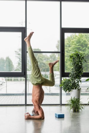 hombre de pelo gris haciendo soporte de cabeza apoyado cerca de bloque de espuma de yoga 