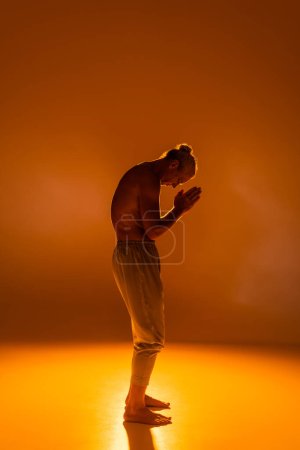 full length of shirtless man in pants practicing yoga with praying hands on orange background 