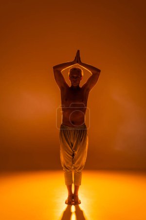 longitud completa del hombre sin camisa en pantalones de pie en pose guerrera sobre fondo naranja 