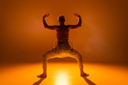 vista trasera del hombre sin camisa practicando la pose de diosa yoga sobre fondo naranja 