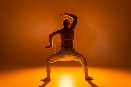 back view of shirtless man practicing goddess yoga pose and gesturing on orange background 
