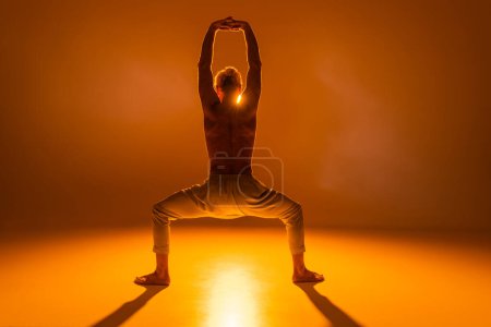 back view of shirtless man practicing goddess yoga pose with raised arms on orange background 