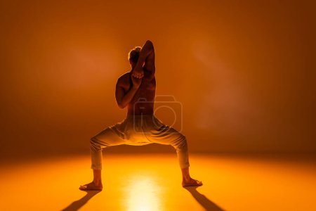 back view of shirtless man practicing goddess yoga pose with clenched hands behind back on orange background 