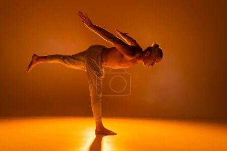 full length of shirtless man in pants doing warrior yoga pose on orange 