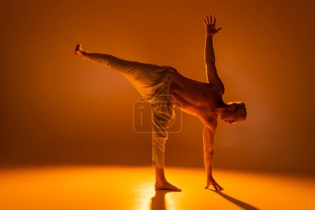 longitud completa de hombre sin camisa en pantalones haciendo media luna pose de yoga en marrón 
