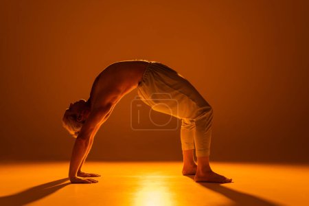 longitud completa de hombre sin camisa en pantalones haciendo pose rueda yoga en marrón 