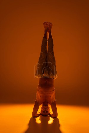 full length of shirtless man doing supported headstand yoga pose on brown 