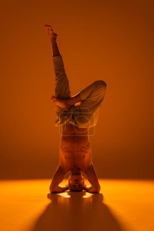 volle Länge des hemdlosen Mannes macht Yoga-Kopfstand-Pose auf braun und orange 