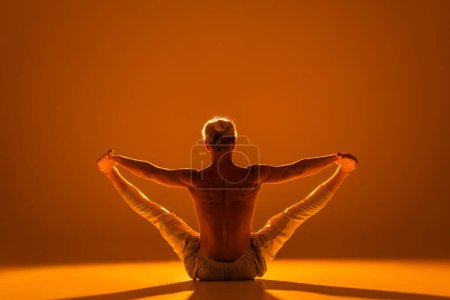 vista posterior del hombre sin camisa haciendo manos sentadas a los dedos de los pies postura de yoga en marrón 