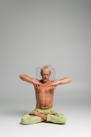 Hemdloser Mann mit geschlossenen Augen sitzt in Lotus-Pose auf grauem Hintergrund 