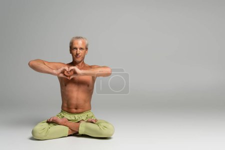 shirtless man in green pants sitting in lotus pose and showing heart sign on grey background 