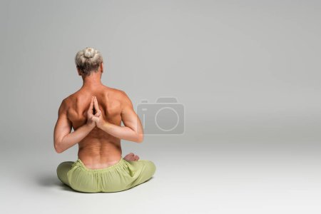 back view of shirtless man in green pants sitting in lotus pose and doing anjali mudra behind back on grey background 