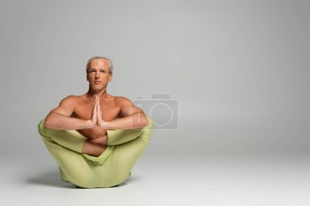 shirtless man in pants sitting in womb embryo pose and doing anjali mudra on grey 
