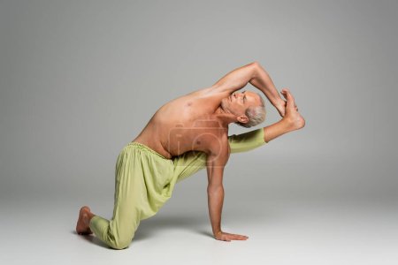 shirtless man in pants doing compass yoga pose on grey background 