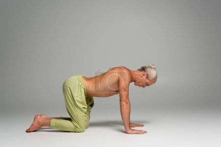 longitud completa de hombre descalzo en pantalones haciendo pose de yoga de vaca en gris 