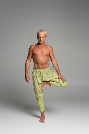 full length of barefoot man in pants standing on one leg in balance yoga pose on grey 