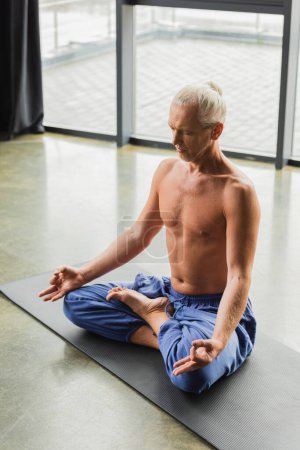 grey haired man in blue pants sitting in lotus pose and doing gyan mudra in yoga studio 