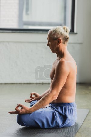 Foto de Side view of grey haired man in blue pants sitting in lotus pose and doing gyan mudra - Imagen libre de derechos