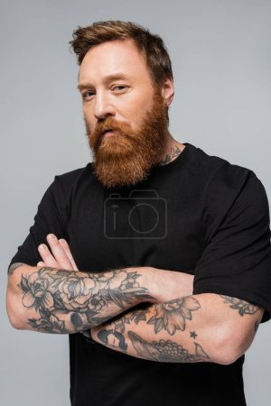 serious tattooed man in black t-shirt standing with crossed arms and looking at camera isolated on grey