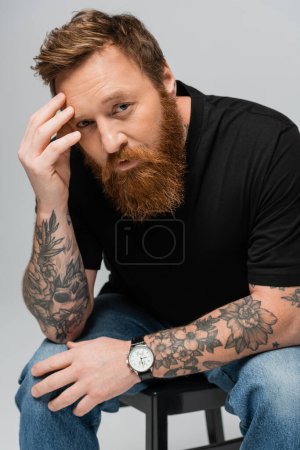 Foto de Hombre tatuado pensativo en camiseta negra y jeans tocando la frente y mirando a la cámara aislada en gris - Imagen libre de derechos