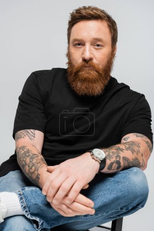 Téléchargez les photos : Homme barbu tatoué en t-shirt noir et jeans assis et regardant la caméra isolé sur gris - en image libre de droit