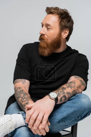 trendy bearded man in black t-shirt and jeans sitting on chair and looking away isolated on grey