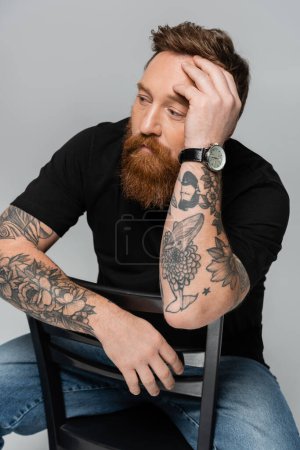 Foto de Hombre tatuado pensativo sentado en la silla con la mano cerca de la cabeza y mirando hacia otro lado aislado en gris - Imagen libre de derechos