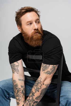 Foto de Hombre tatuado reflexivo en camiseta negra sentado en la silla mientras mira hacia otro lado aislado en gris - Imagen libre de derechos