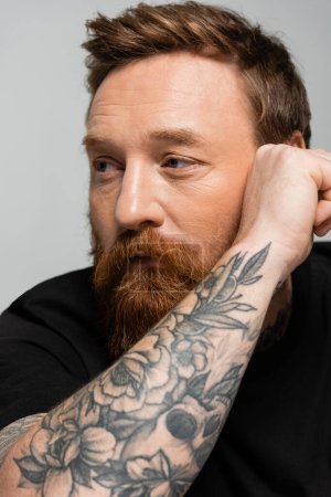 Téléchargez les photos : Portrait d'un homme barbu réfléchi tenant la main tatouée près du visage et regardant loin isolé sur le gris - en image libre de droit