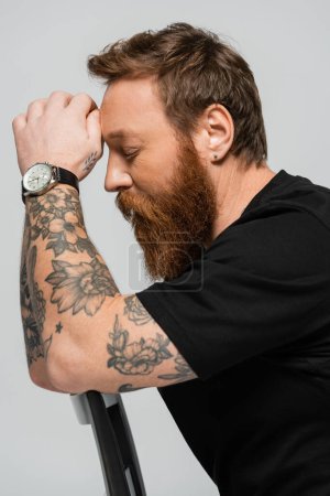 Photo for Side view of tired tattooed man in black t-shirt and wristwatch sitting with closed eyes isolated on grey - Royalty Free Image