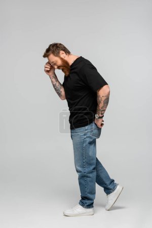 Téléchargez les photos : Side view of smiling man in black t-shirt and jeans touching forehead while smiling on grey background - en image libre de droit