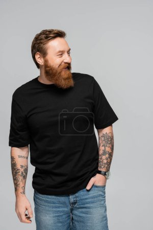 fröhlicher bärtiger Mann im schwarzen T-Shirt, der mit der Hand in der Jeanstasche steht und isoliert auf grau blickt