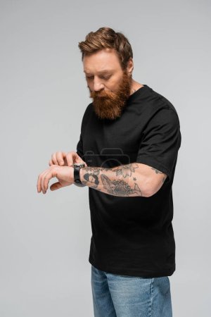 hombre tatuado y barbudo en camiseta negra mirando reloj de pulsera aislado en gris