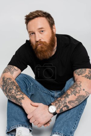 serious bearded man in black t-shirt and jeans looking at camera while sitting on grey background