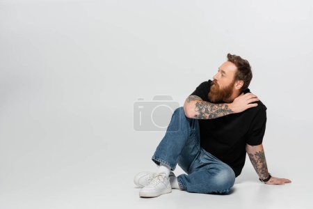 Foto de Longitud completa del hombre tatuado en pantalones vaqueros azules y zapatillas blancas sentado y mirando hacia otro lado sobre fondo gris - Imagen libre de derechos