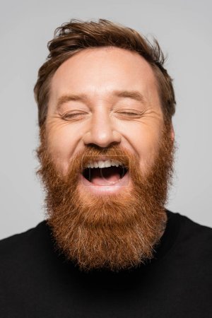 portrait of excited bearded man laughing with closed eyes isolated on grey