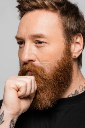 portrait of pensive bearded man holding hand near face and looking away isolated on grey