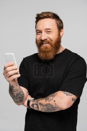 fröhlicher bärtiger Mann im schwarzen T-Shirt lächelt in die Kamera, während er sein Handy isoliert auf grau hält